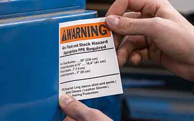 A person applies a bright orange arc flash warning label to a machine.