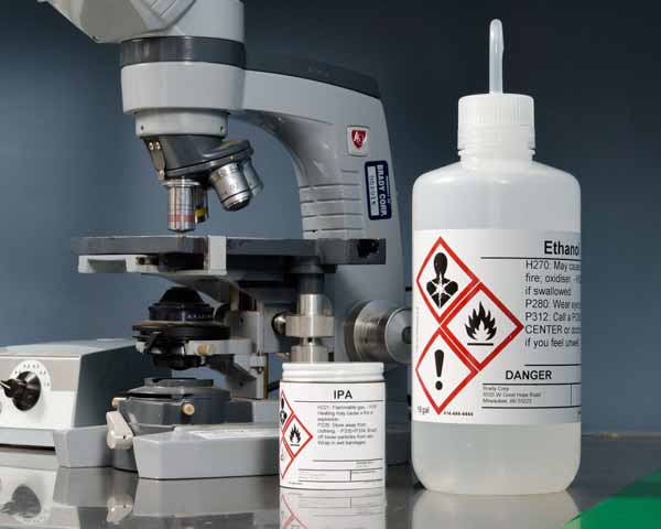 A microscope positioned alongside bottles of Isopropyl Alcohol (IPA) and Ethanol, each bearing Globally Harmonized System (GHS) labels for safety and chemical identification.
