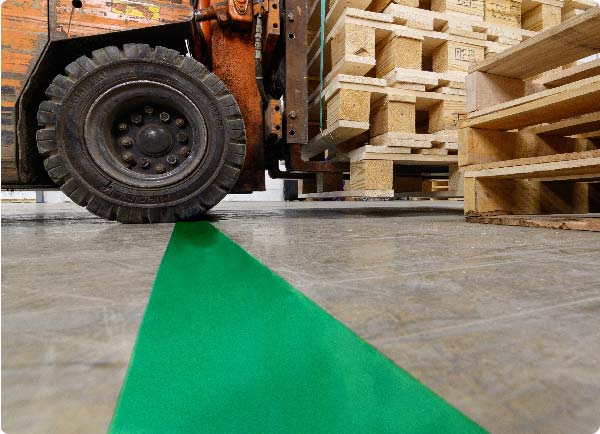 A large forklift is driving over a line of green ToughStripe Max floor tape. The edges of the tape are undamaged by the forklift's heavy-duty wheels.