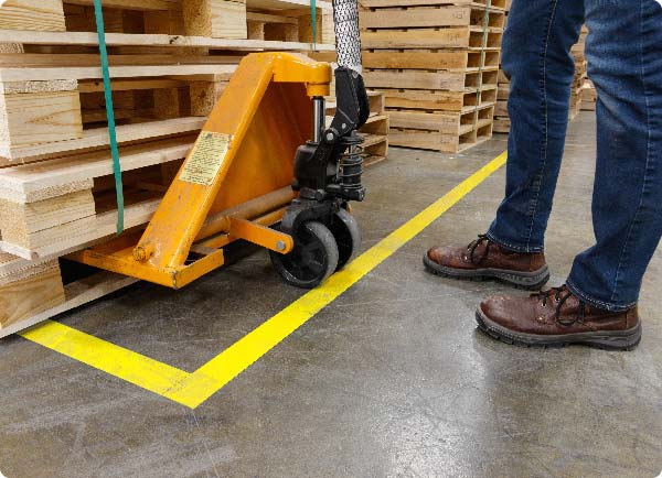 Bright yellow ToughStripe floor tape is applied to a warehouse floor. Its position indicates where a pallet jack operator should deposit a stack of materials.