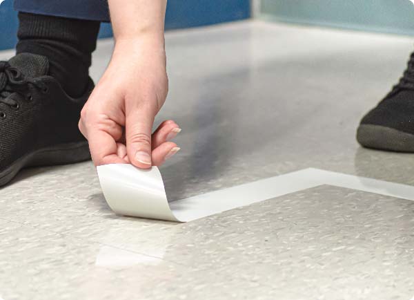 A person is using their hands to remove ToughStripe Vinyl floor tape from vinyl composition tile. The tape is coming off cleanly in one piece.