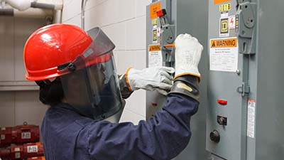 A woman wearing a safety mask inspects a breaker box.