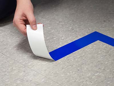A person applies blue area marking tape to synthetic tile floor.