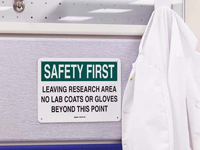 A lab coat hangs on a wall next to a sign that says 'Safety first. Leaving research area. No lab coats or gloves beyond this point.'