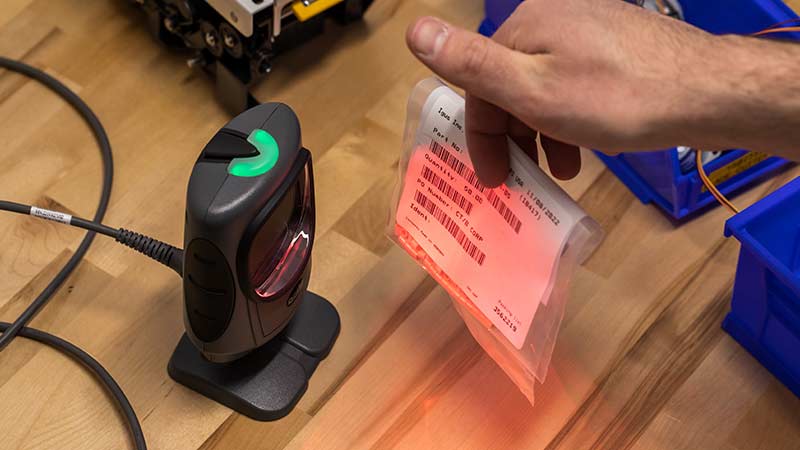A worker scans a barcode using a fixed barcode scanner on a work bench.