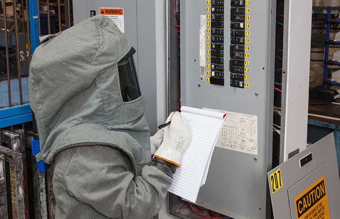 An engineer wearing a full-body safety suit performing an audit. He is standing in front a breaker box and is holding a clipboard.