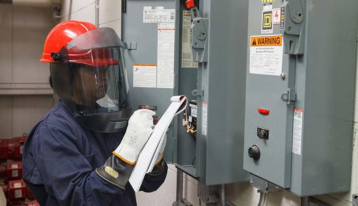 A worker in full protective gear working though a checklist at a panel with arc flash pumps.