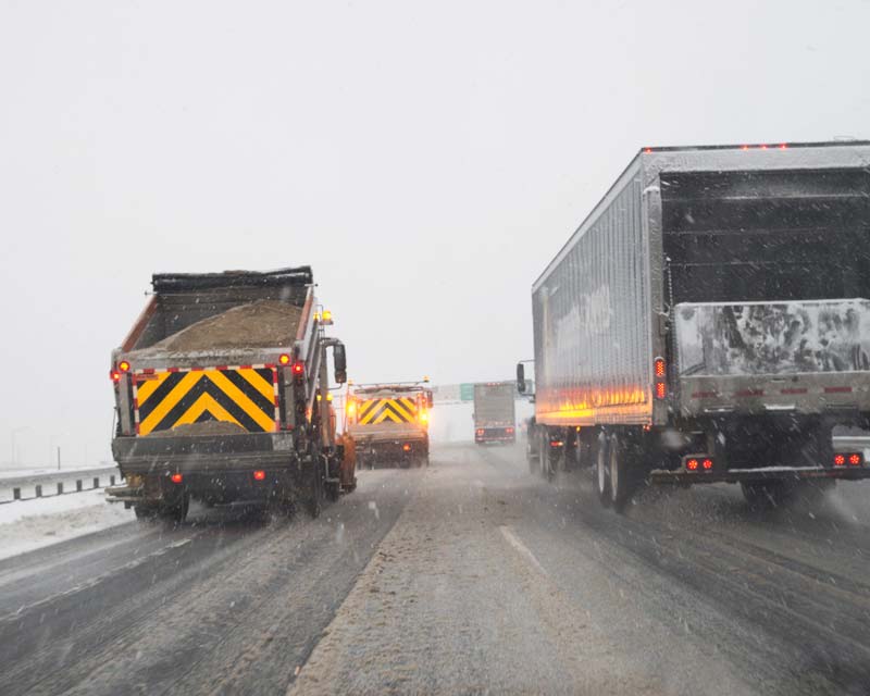 Two DOT trucks driving on a highway in snowy conditions showing usage of approved reflective tape.