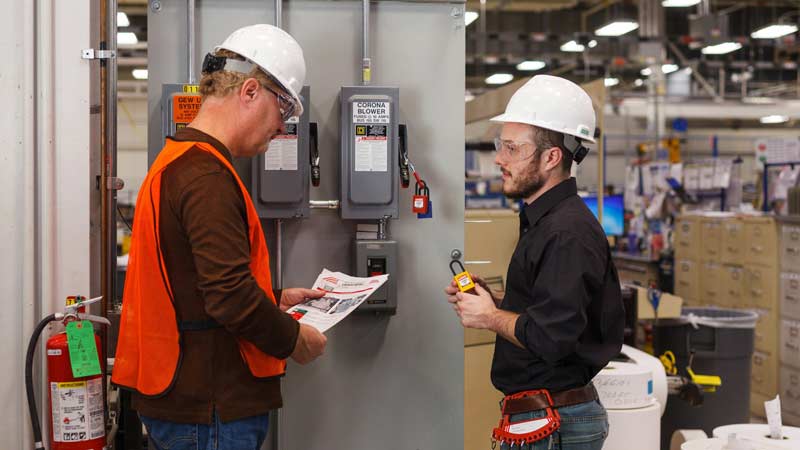 Two workers review the lockout tagout procedure for an electrical panel in a warehouse setting.