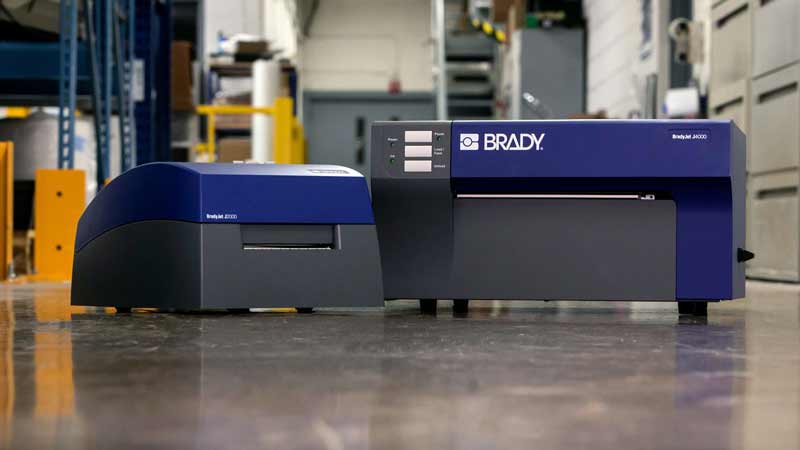 Two Brady inkjet label printers sit on a concrete floor in a warehouse setting.