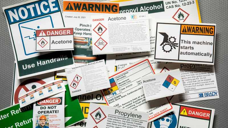 A gray notice board filled with a variety of safety signage and label examples.