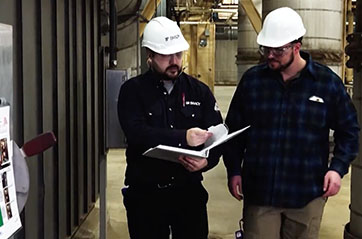 Two men walking through a manufacturing facility. One is wearing a hardhat with a Brady logo, and he is pointing to a binder.