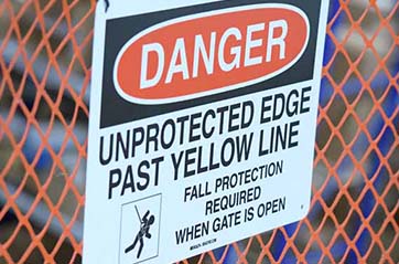 A sign on a construction fence. It says "DANGER, UNPROTECTED EDGE PAST YELLOW LINE."