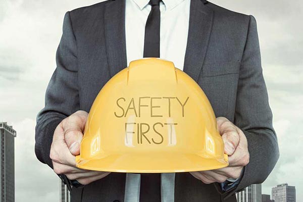 A person dressed in business attire holds out a hard hat that has the words “safety first” written on it.