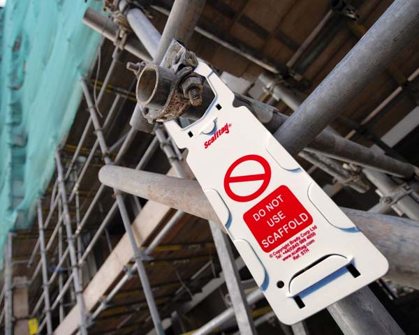 A white and red scaffold tag hangs on the outside scaffolding of an industrial building.