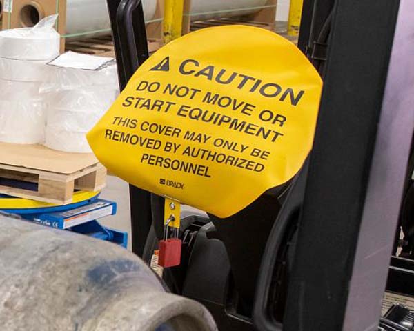 A forklift is shown in a warehouse with a steering wheel safety cover on it that says 'Caution - Do Not Move Or Start Equipment - This Cover May Only Be Removed By Authorized Personnel.'