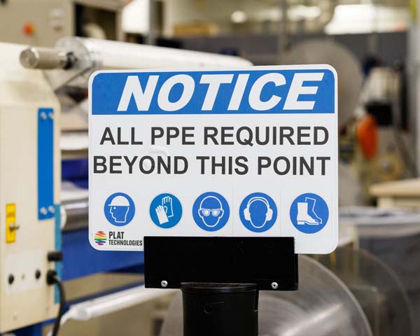 A white a blue OSHA PPE Sign that says 'Notice - All PPE Required Beyond This Point' is displayed on a warehouse floor.