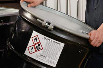 A person lifting a barrel that's labeled with GHS warnings.