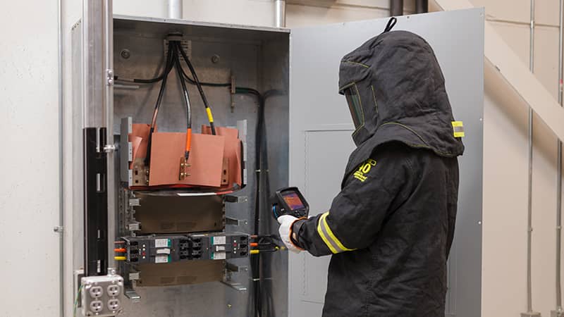 A worker wearing PPE uses an infrared camera on an electrical panel in a warehouse setting.