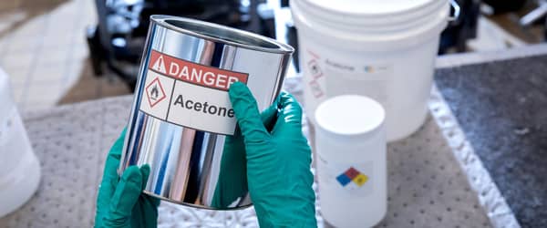 A worker wearing latex glove PPE inspects a container with a chemical labels stating they should be aware of the danger of the acetone contents.