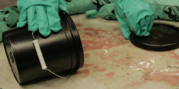 A container containing an unknown hazardous chemical is shown spilled on a concrete floor while a worker wearing PPE begins the cleaning process.