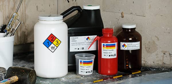 Three different sized secondary containers with proper HazCom labeling are sitting on display in an industrial environment.