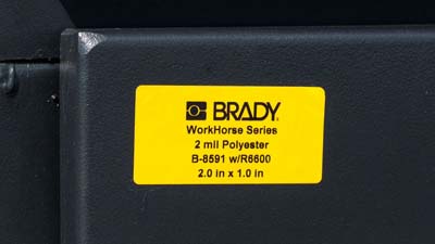 A yellow Brady all-weather polyester label with printed black text applied to a piece of equipment.