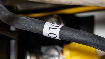 A white self-extinguishing tedlar wrap around label with black text applied around a black wire of an electrical box.